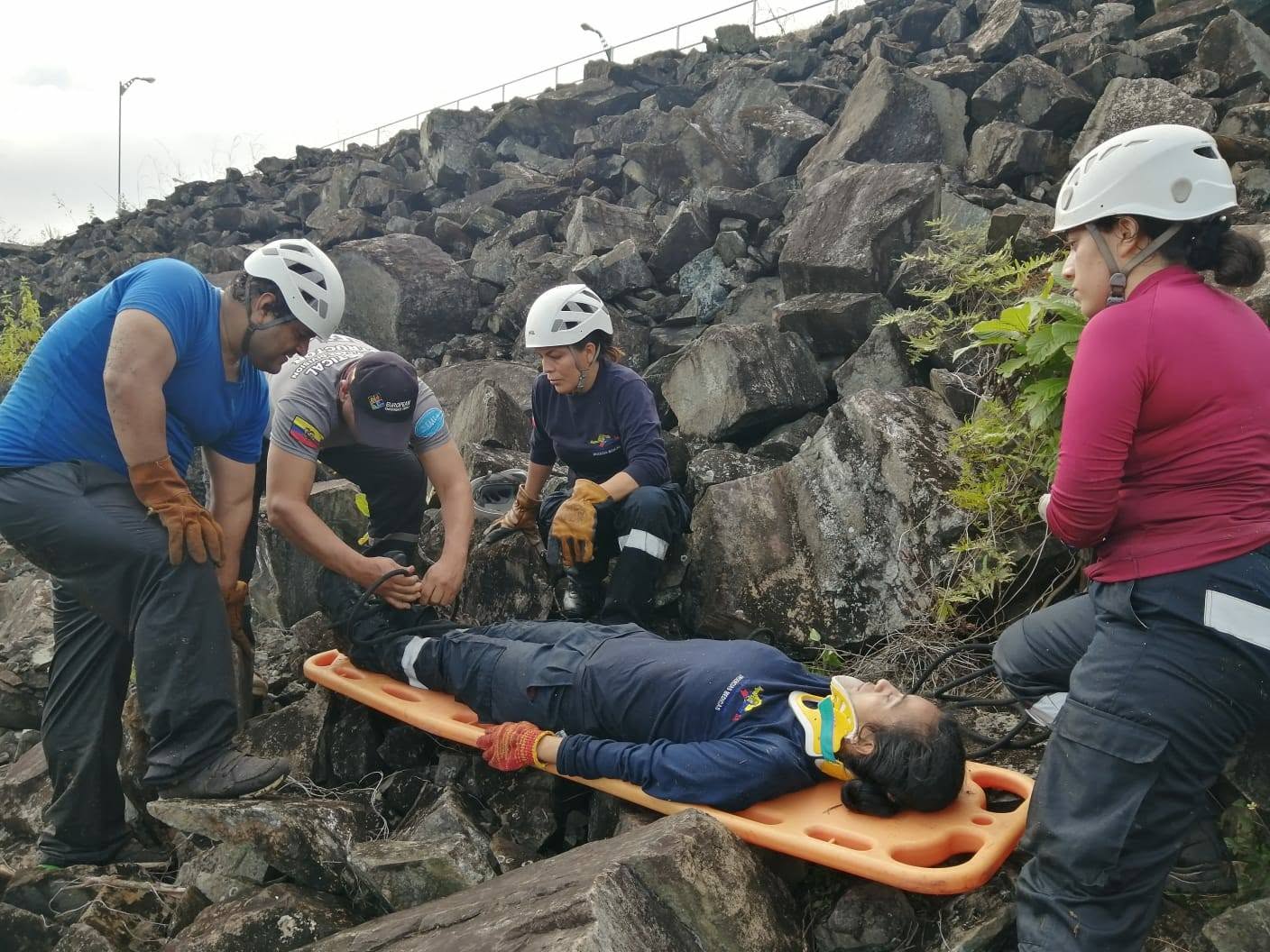 Members of an EEC rescue team wearing helmets and protective gloves prepare to move a person on an orange stretcher across rocky ground.